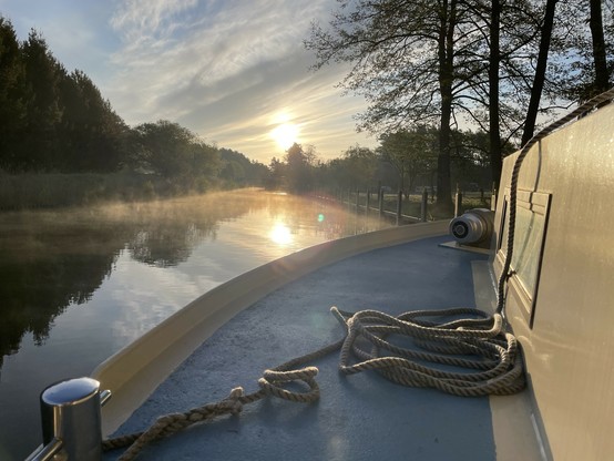 Blick auf den Sonnenaufgang vom Bug eines Bootes auf einem ruhigen Fluss mit vom Wasser aufsteigendem Nebel und Bäumen entlang des Ufers.