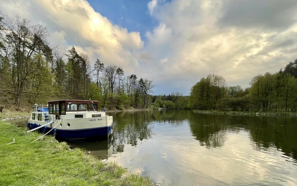 Ein auf einem ruhigen Fluss vertäutes Boot mit Spiegelungen von Wolken und Bäumen auf dem Wasser, flankiert von Grünflächen und einem Waldgebiet.