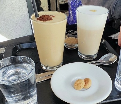 Ein Tablett mit drei sichtbaren Gläsern. Vorne links ist ein Wasserglas. Hinten links ist ein Glas Sorbetto. Hinten rechts ein Cappuccino (ausnahmsweise im Glas für den Mann). Vorne ein  Teller mit zwei Amarettini.