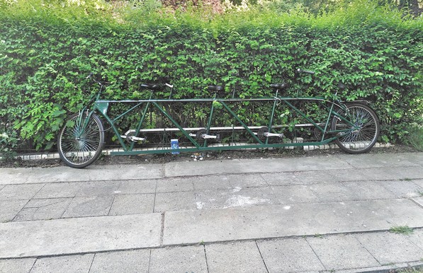 At the top there is a green hedge and the the bottom grey square and rectangular paving slabs. In the middle of the image is a dark green bicycle with four seats. 