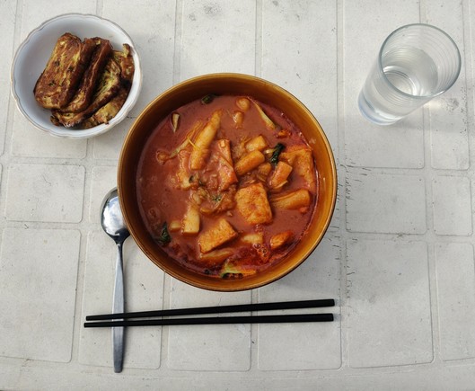 On a white table is a big bowl with tteokbokki, a small bowl with fried slices of a rolled omelette, a glass of water, chopsticks, and a spoon.