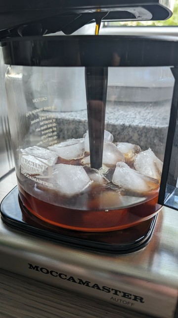An image of a silver moccamaster coffee maker. The pot is filled with ice cubes and fresh coffee is brewed onto that.