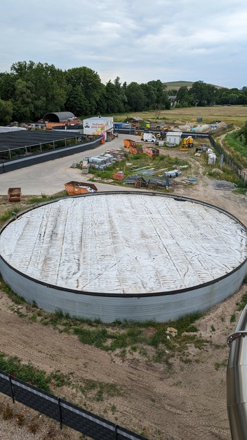 A photo of a very large water reservoir covered with a large light grey tarpaulin. The folds and dirt on the foil make the structure look like an oven cheese with a diameter of 20 metres.