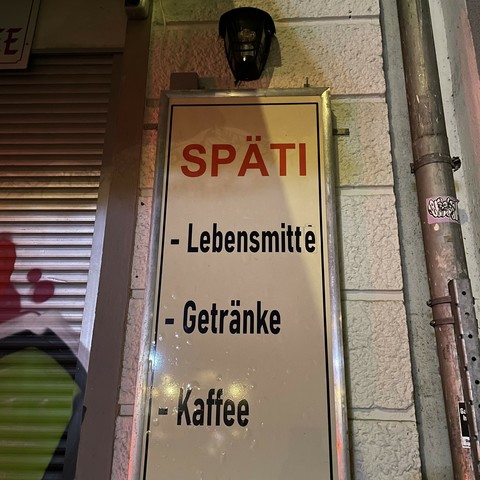 Ein Metallschild an einem Späti in Berlin, neben einem Fenster mit heruntergelassenen Rollläden. Unter der Überschrift Späti finden sich die Punkte „Getränke“ und „Kaffee“, an erster Stelle aber befindet sich der Punkt „Lebensmitte“ (sic). 

Über dem Schild befindet sich eine Warnlampe eines Alarmsystems, daneben eine Regenrinne des Nachbargebäudes. 