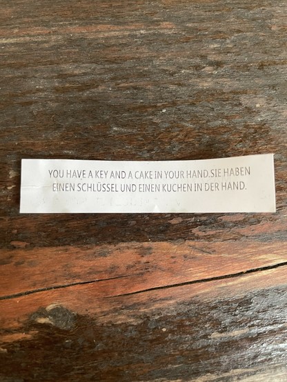 Ein Zettel aus einem Glückskeks der auf einer Holzoberfläche liegt. Darauf steht geschrieben: 