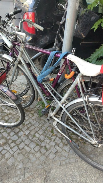 Ein Fahrrad mit einem blauen Schloss an einen Pfahl angeschlossen, dahinter ein anderes Fahrrad, das dadurch eingeklemmt ist. 