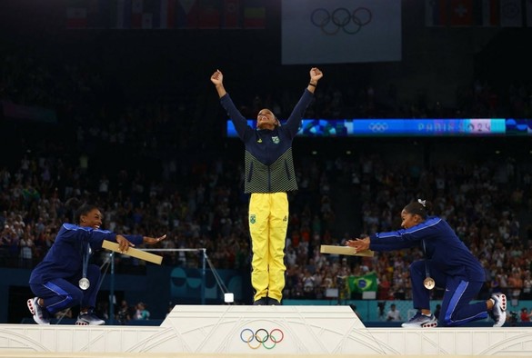 Rebeca Andrade stepping up on the podium to receive the gold medal, Simone Biles and Jordan Chiles reverencing her