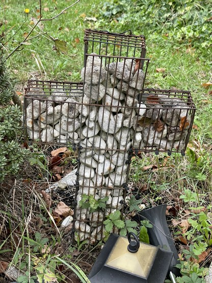 Ein mit kleinen Steinen gefülltes Kreuz aus Drahtgeflecht ist annehmen kleinen Urnengrab als Grabstein aufgestellt. Drumherum ist Erde und Gras, kleines Friedhofsgrünzeug. Das Drahtgeflecht ist kaputt, die Steine sind nach unten gesackt und füllen das Kreuz nicht mehr aus. 