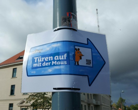 Ein blauer Pfeil mit Mauslogo auf einem weißen Zettel an einem Laternenmast zeigt in Richtung des Funkmuseums, wo der Maustag stattfindet. Im Hintergrund ein Haus, ein Antennenmast und dunkle Wolken