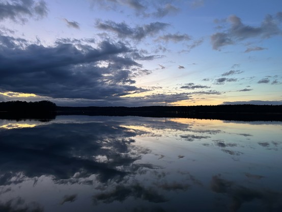 Blick auf einen See, in der Mitte des Bildes ist das Ufer zu sehen. Die Sonne ist gerade untergegangen.
Im Vordergrund Wasser in dem sich der wolkige Himmel des oberen Teil des Bildes spiegelt