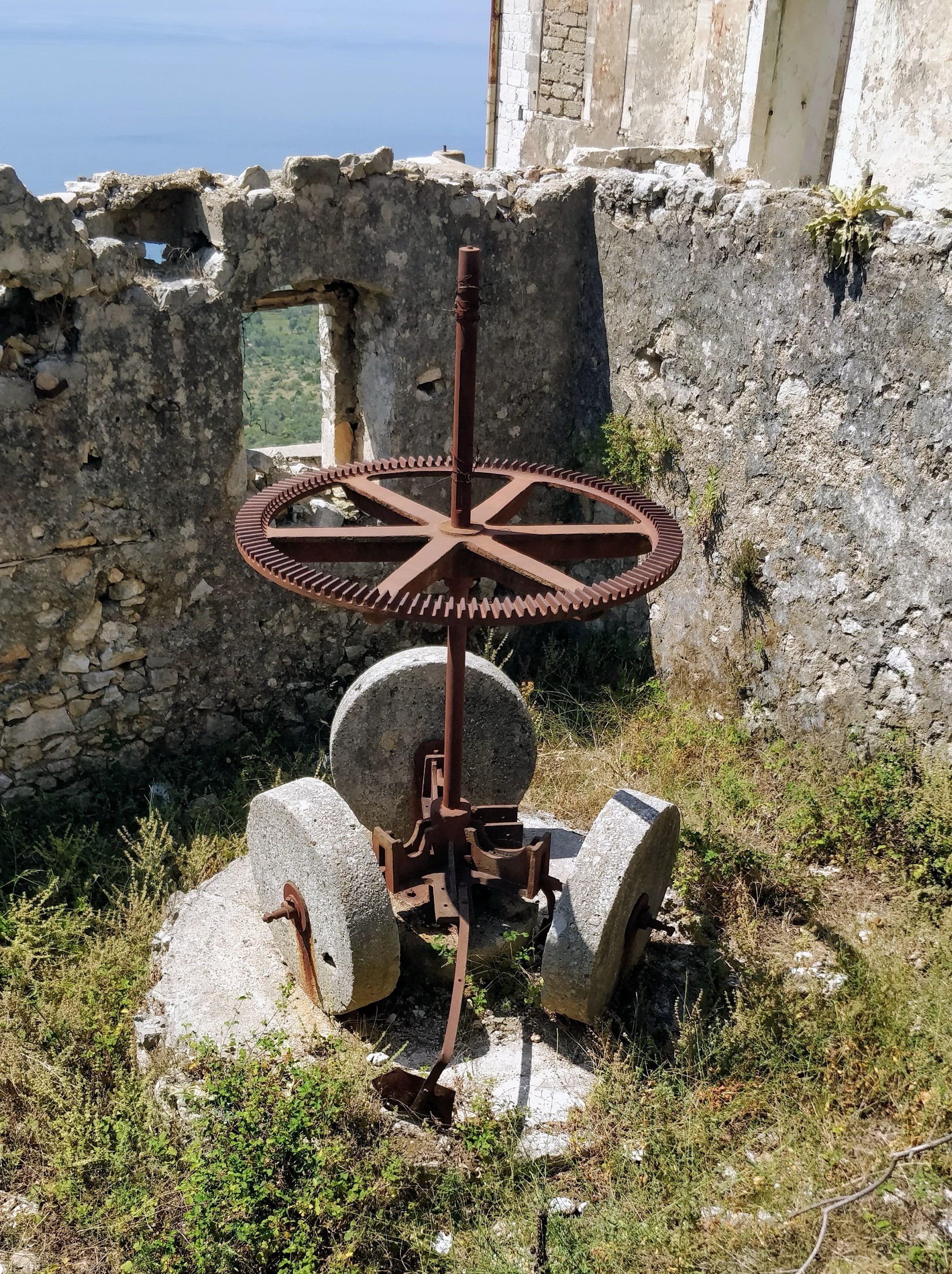 Ein alter Mühlstein mit einem Zahnrad auf der Antriebswelle steht inmitten von Grün in einem zerfallenden Gebäude. Im Hintergrund kann man das Meer sehen. 