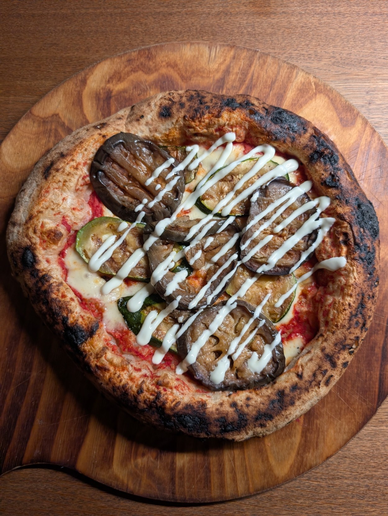 Ein Bild einer neapolitanischen Pizza auf einem Holzbrett. Der Rand ist luftig aufgegangen. Fie Pizza ist mit Tomatensoße, Fior di Latte und Antipasti (Aubergine, Zucchini) belegt und danach mit Aioli garniert.