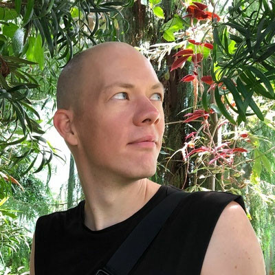 Alex Feyerke is pictured in full colour here, looking off thoughtfully and smiling in front of lush, flowering greenery.